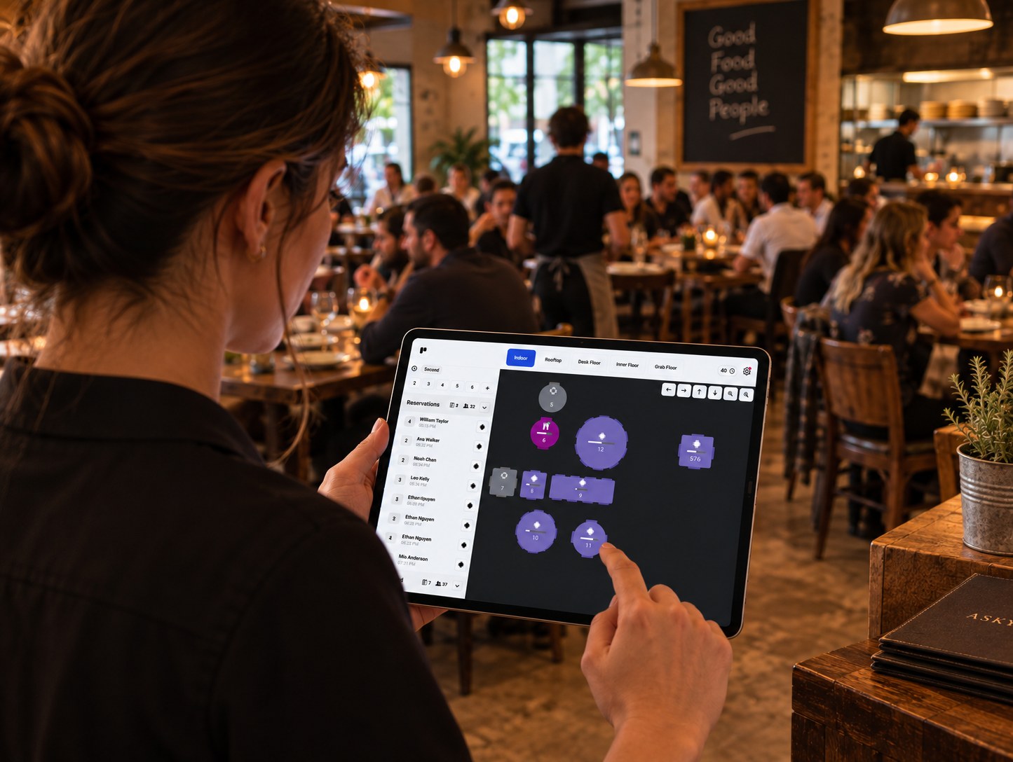 A staff member checking the live floor view on a tablet in a busy dining room, tables shown by colour and status